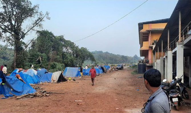 Blue-tarp tents of railway labourers stand along a dirt path just beside the concrete pillars of Chandor station.