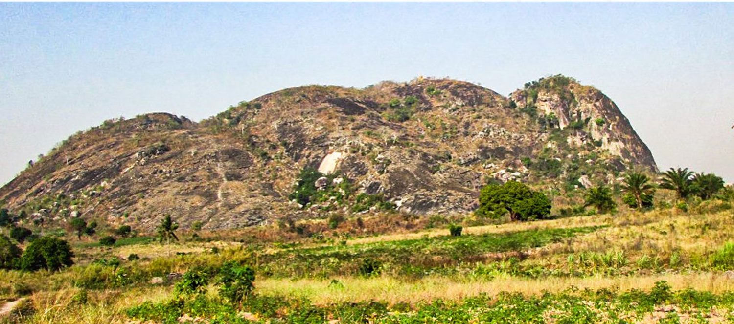 A rounded granite hill rising above dry grassland and scattered palms under a pale sky